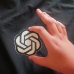 A hand holding a circular OpenAI sticker showing the OpenAI logo