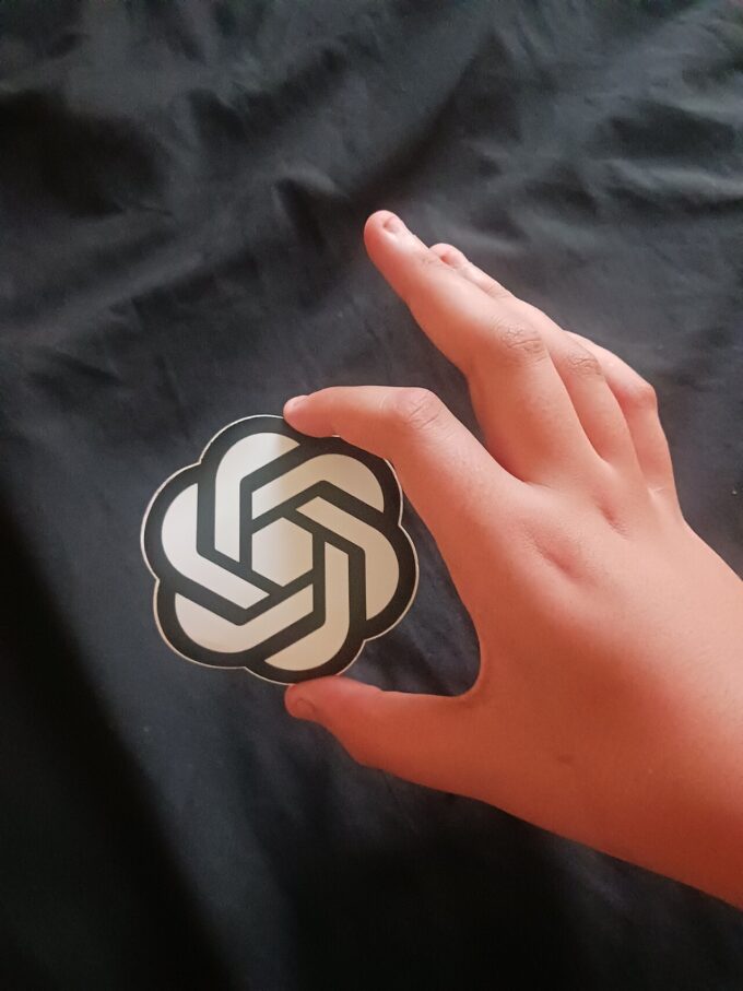 A hand holding a circular OpenAI sticker showing the OpenAI logo